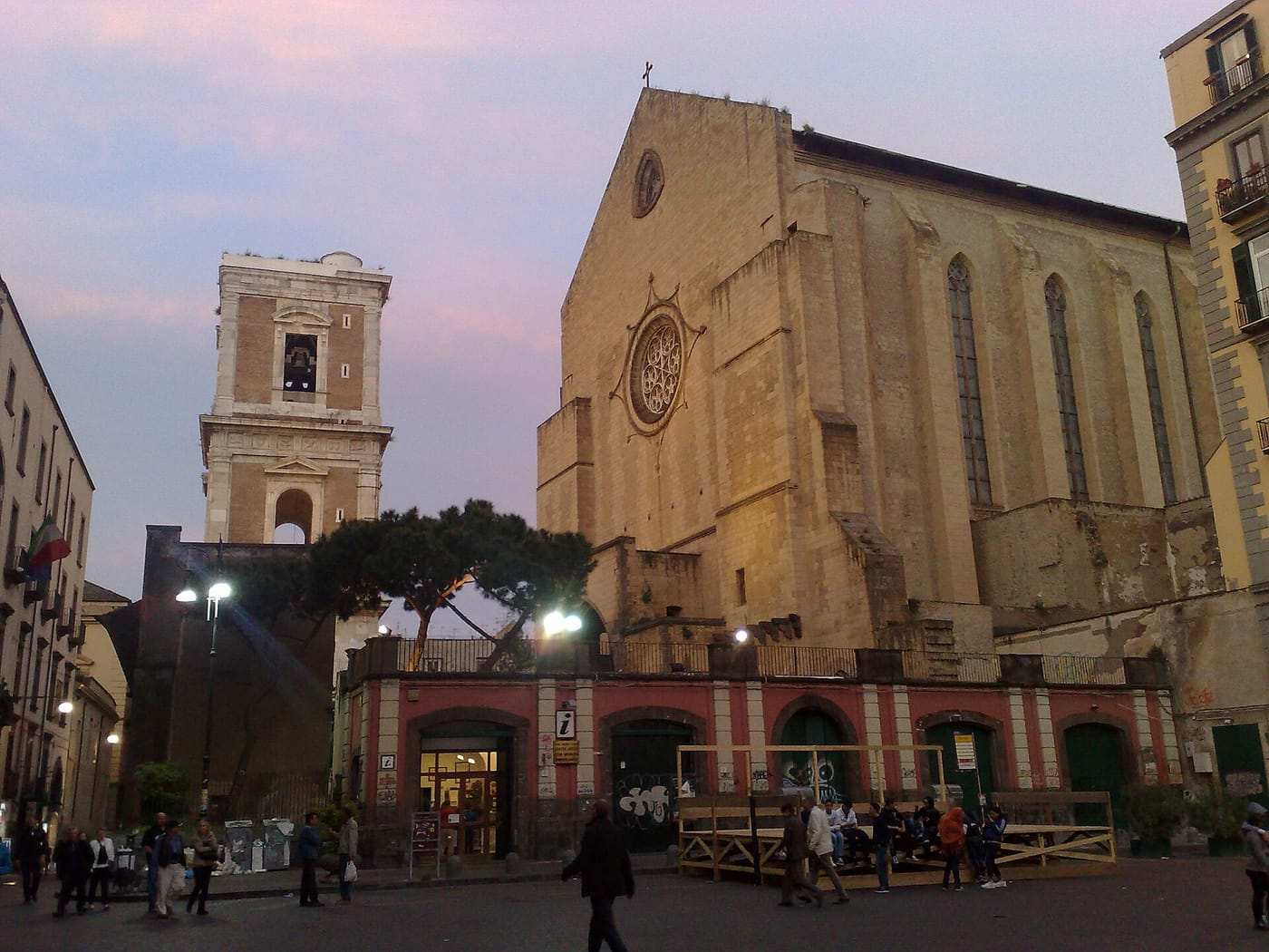 La basilica gotica di Santa Chiara in tufo e il suo campanile al tramonto, con persone che passeggiano davanti.