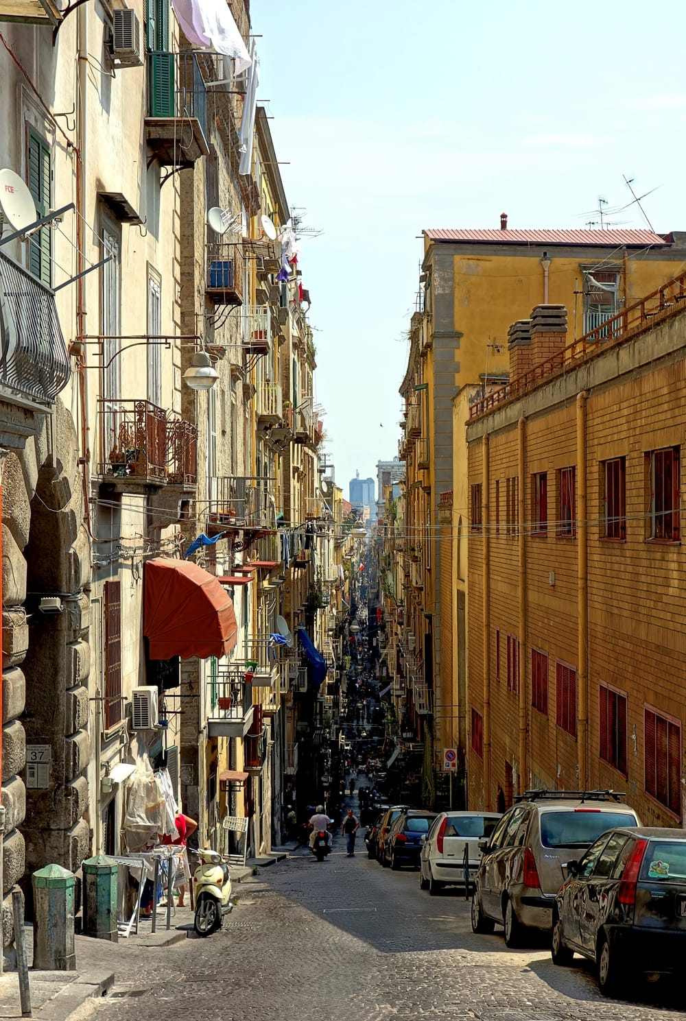Una via stretta e dritta del centro storico tra palazzi alti e colorati con balconi; la strada prosegue fino in fondo, piena di passanti.
