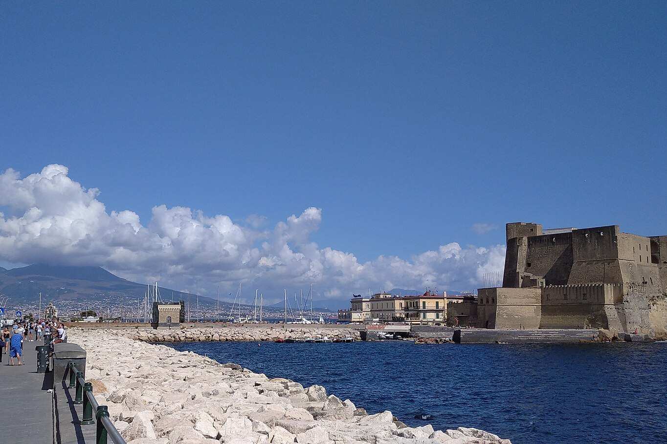 Il lungomare di via Partenope in una giornata di sole: la scogliera bianca lungo la passeggiata, il mare blu e, in fondo, Castel dell'Ovo sull'isolotto.