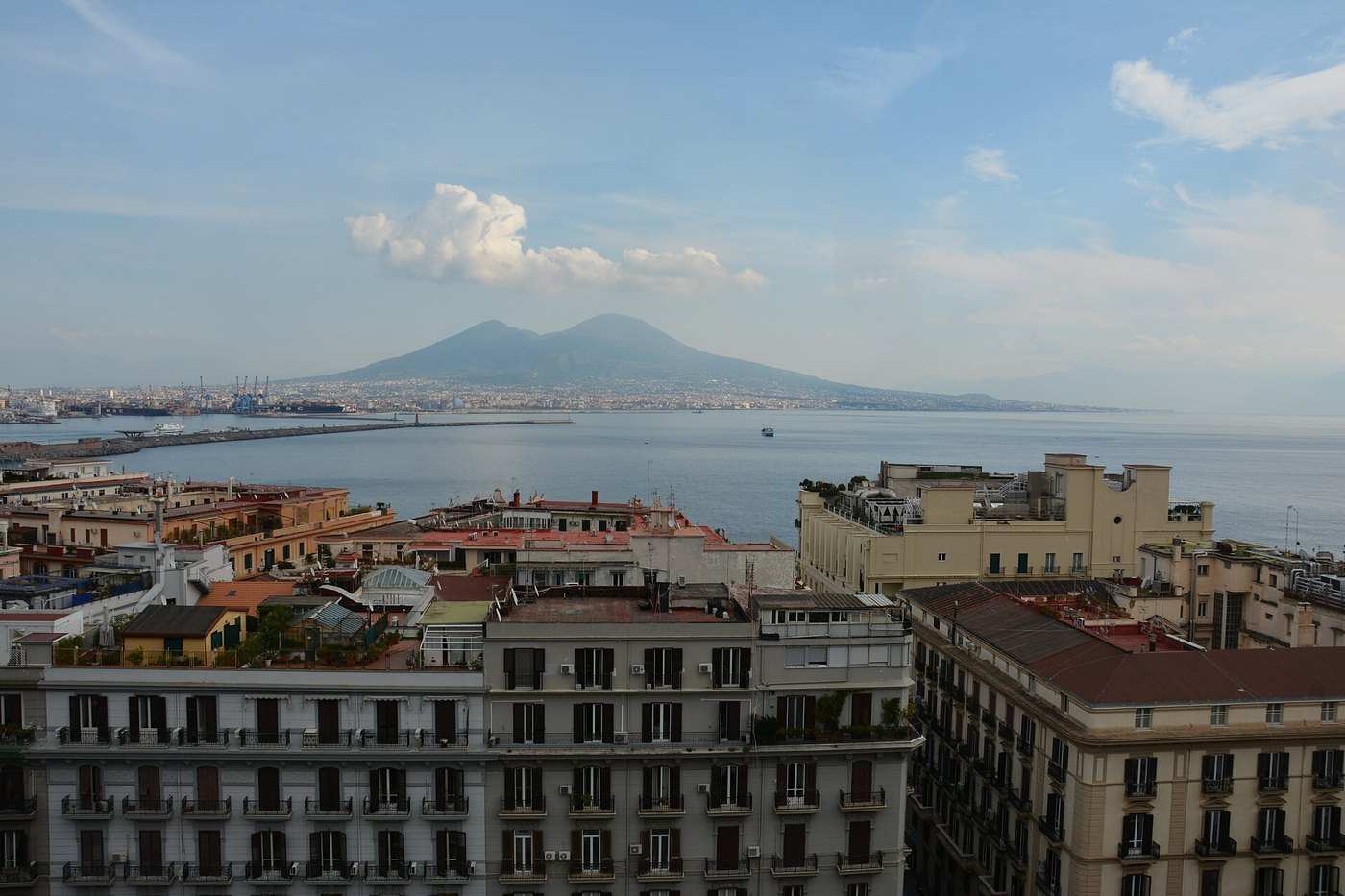 Dall'alto di Monte Echia: in primo piano i tetti e i palazzi di Santa Lucia, poi il mare del golfo e, sullo sfondo, il Vesuvio sotto un cielo con qualche nuvola.